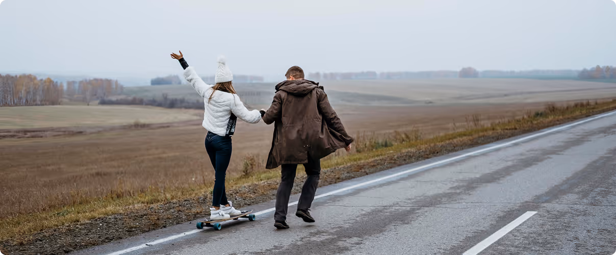 A woman rides a skateboard on a rural road holding hands with a man walking beside her, with open fields and trees in the background under a cloudy sky.