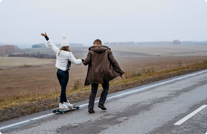 Couple on a rural road with woman riding a longboard and man walking beside her holding hands.