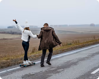 Rear view of a couple holding hands outdoors, with the woman riding a skateboard on an empty rural road.