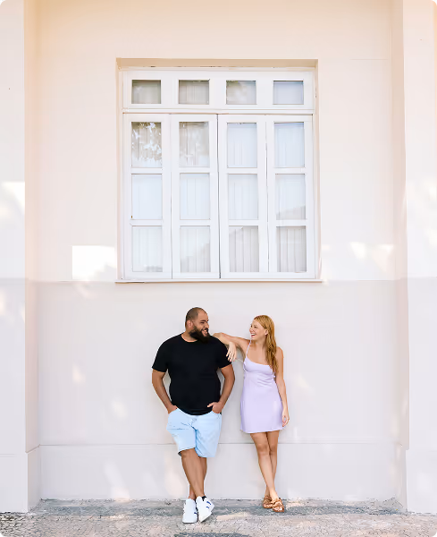 Man in black shirt and light blue shorts and woman in lavender dress standing and smiling in front of a beige wall with a large white window.