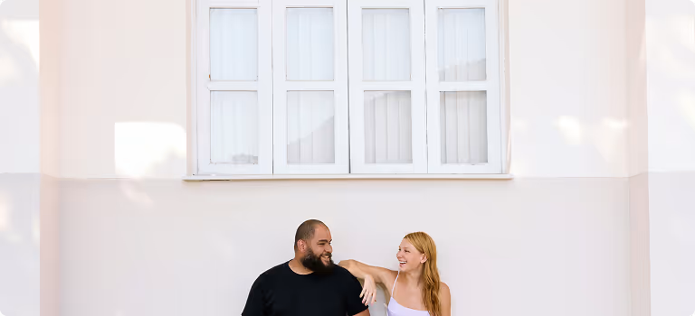 Smiling couple sitting and talking under a large window on a light-colored wall.