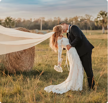 Bride and groom kissing in a grassy field beside a hay bale during sunset, bride holding flowers and veil flowing.