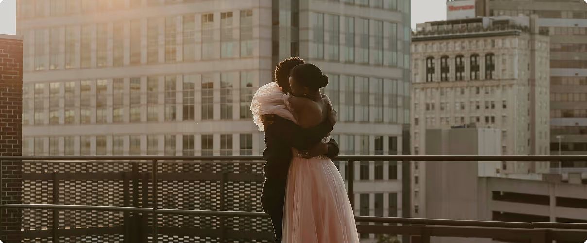 Couple embracing on an outdoor balcony with a cityscape of tall buildings in the background during sunset.