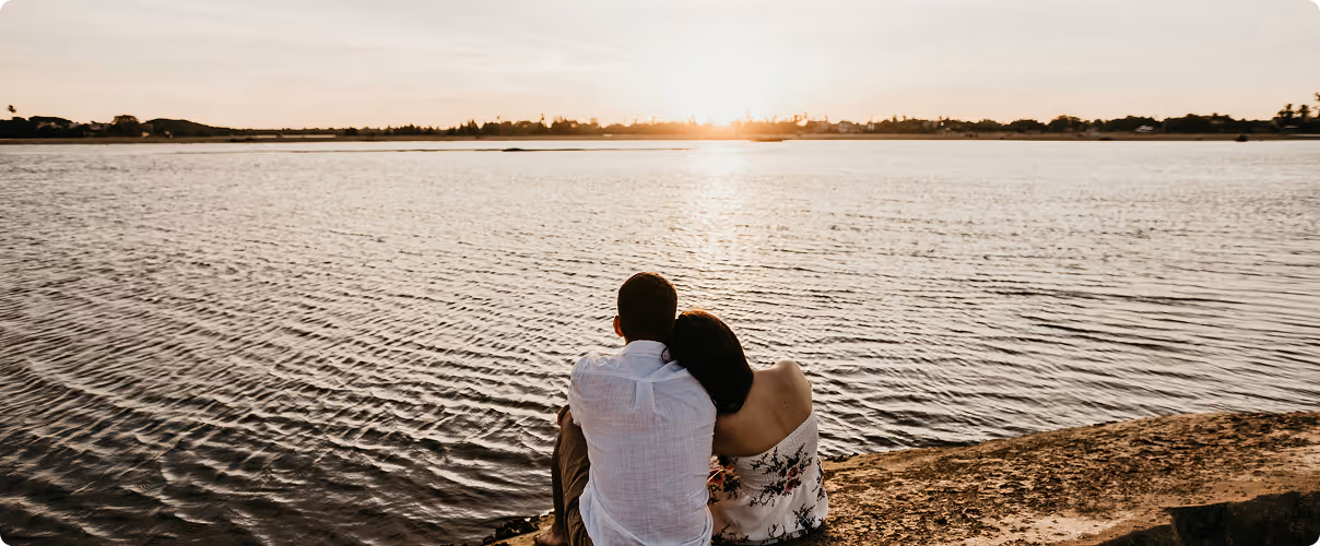 Couple sitting close together by a lakeside at sunset, with the sun low on the horizon over calm water.