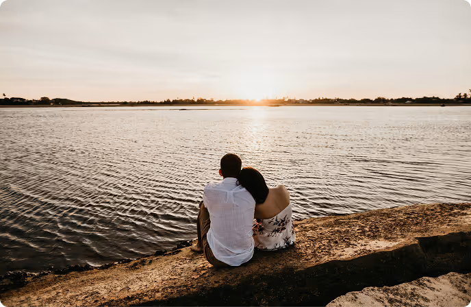 Couple sitting on a sandy shore, facing a calm body of water with the sun setting on the horizon.