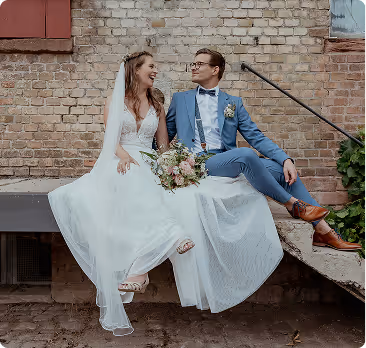 Bride in white dress and groom in blue suit sitting on an outdoor concrete ledge against a brick wall, smiling at each other.
