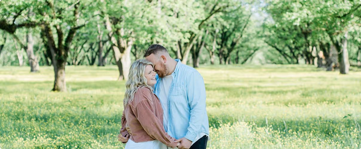 Couple holding hands and smiling intimately in a sunlit green orchard.