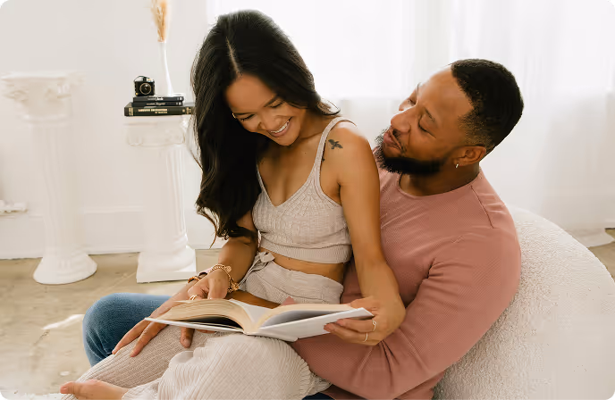 Couple sitting closely on a white chair, the woman reading a book while the man embraces her from behind, both smiling.