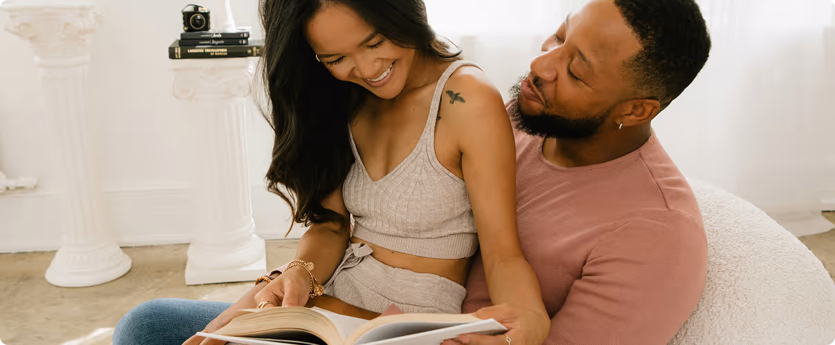 Smiling couple sitting closely on a chair reading a book together in a bright room.