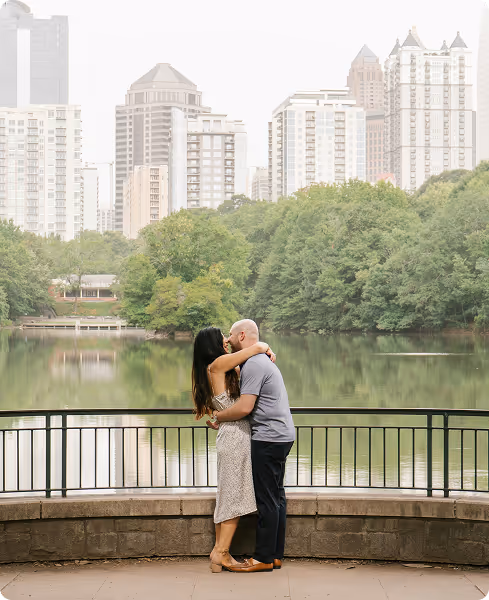 Couple embracing and kissing on a lakeside walkway with city buildings and trees in the background.