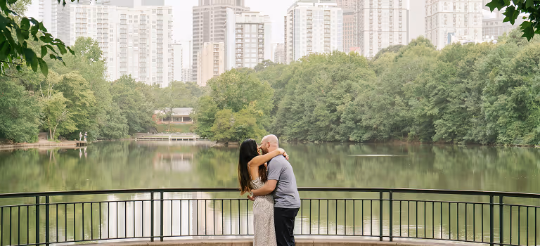 Couple embracing and kissing on a railing overlooking a calm lake with dense trees and city buildings in the background.