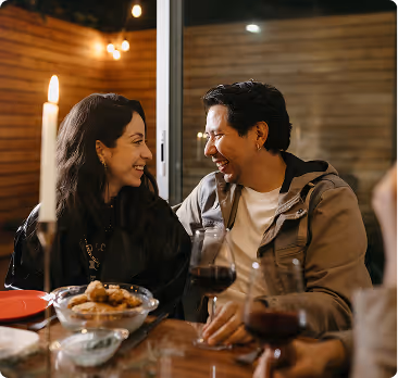Couple sitting at a table indoors, smiling and looking at each other with wine glasses and snacks on the table.