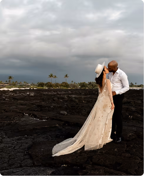 Couple kissing on dark volcanic rock beach under cloudy sky, woman in white lace dress and hat, man in white shirt and black pants.