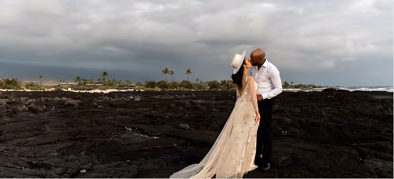 Couple kissing on black volcanic rocks with cloudy sky and palm trees in the background.