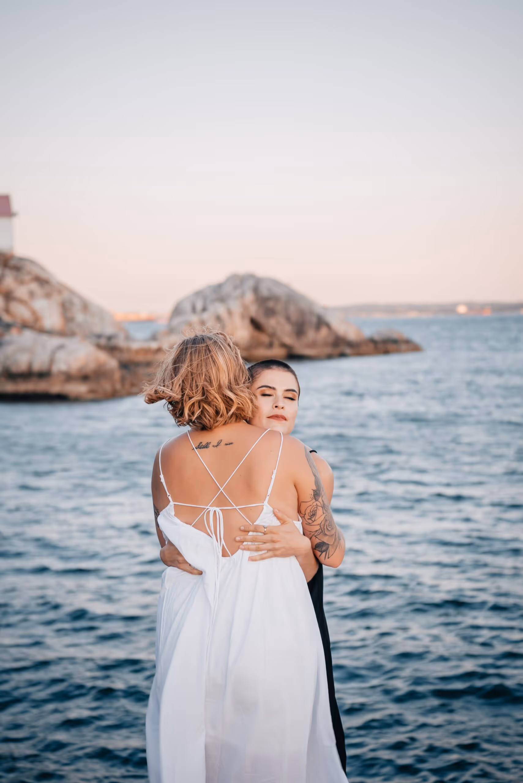 Two women embracing by the sea with rocky shore in the background during sunset.