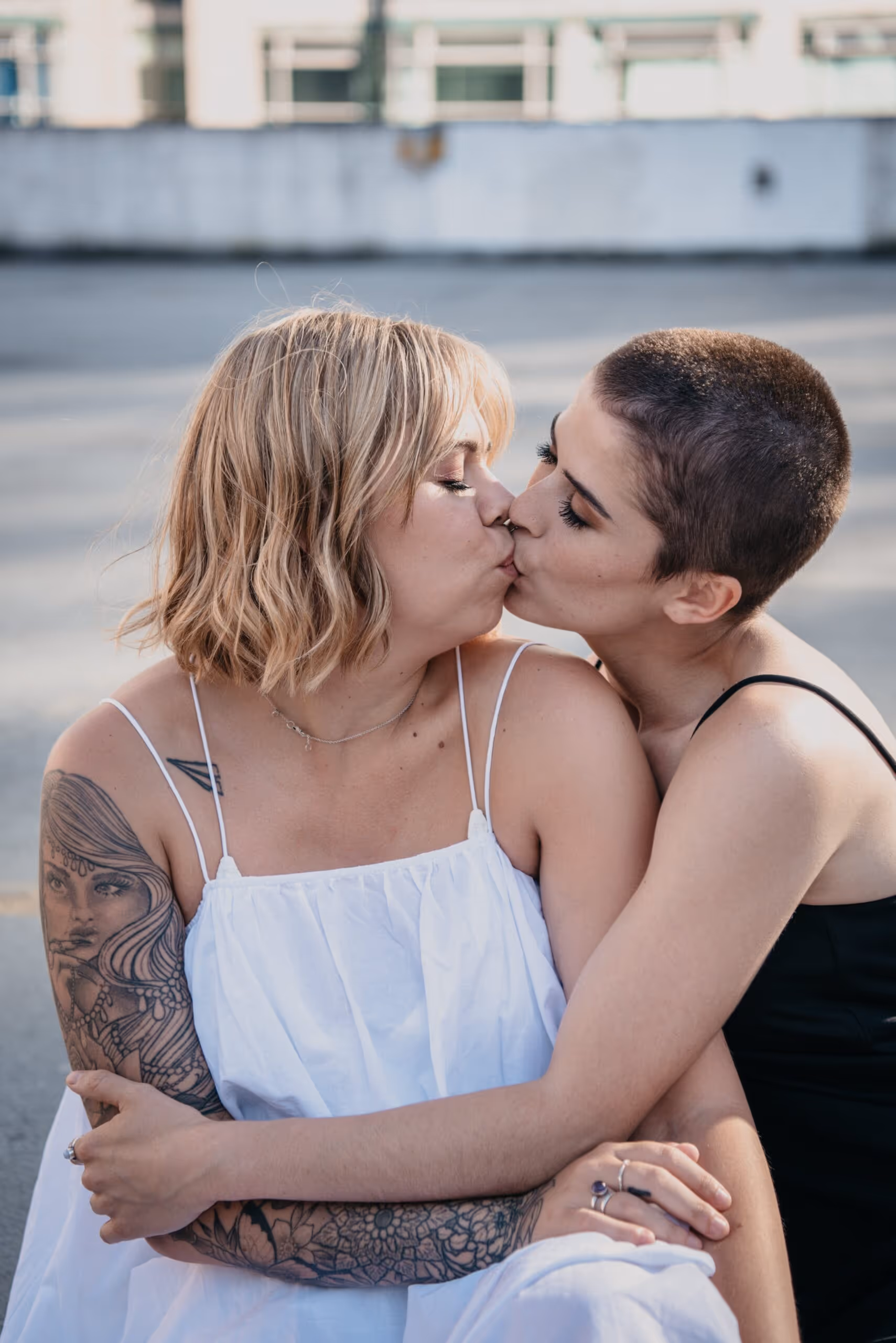 Couple embracing and sharing a kiss outdoors, one with short hair in a black top and the other with a tattooed arm in a white dress.