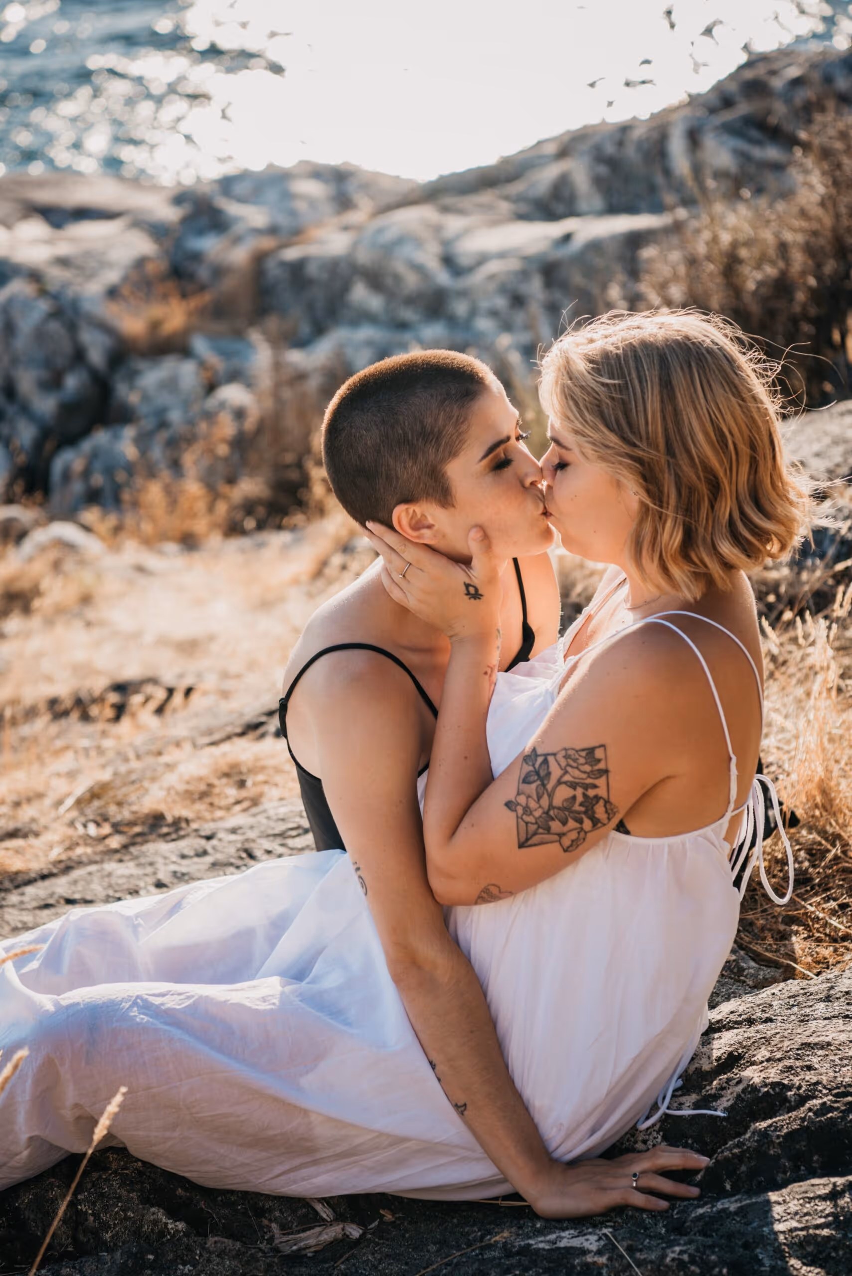 Two women sitting on rocks outdoors sharing a tender kiss, one with short hair and the other with tattoos on her arm.