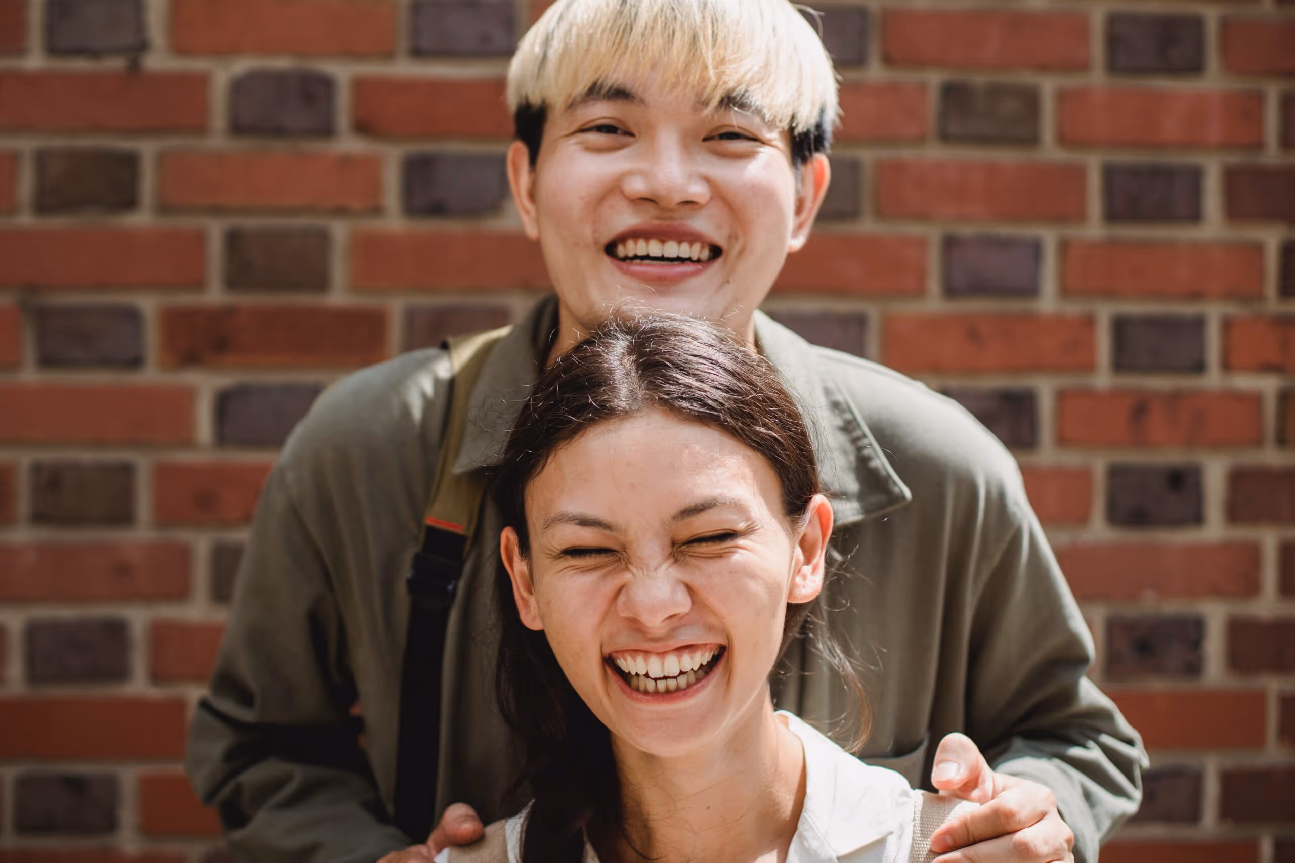 Happy young man with blond hair standing behind a laughing woman with dark hair against a brick wall.