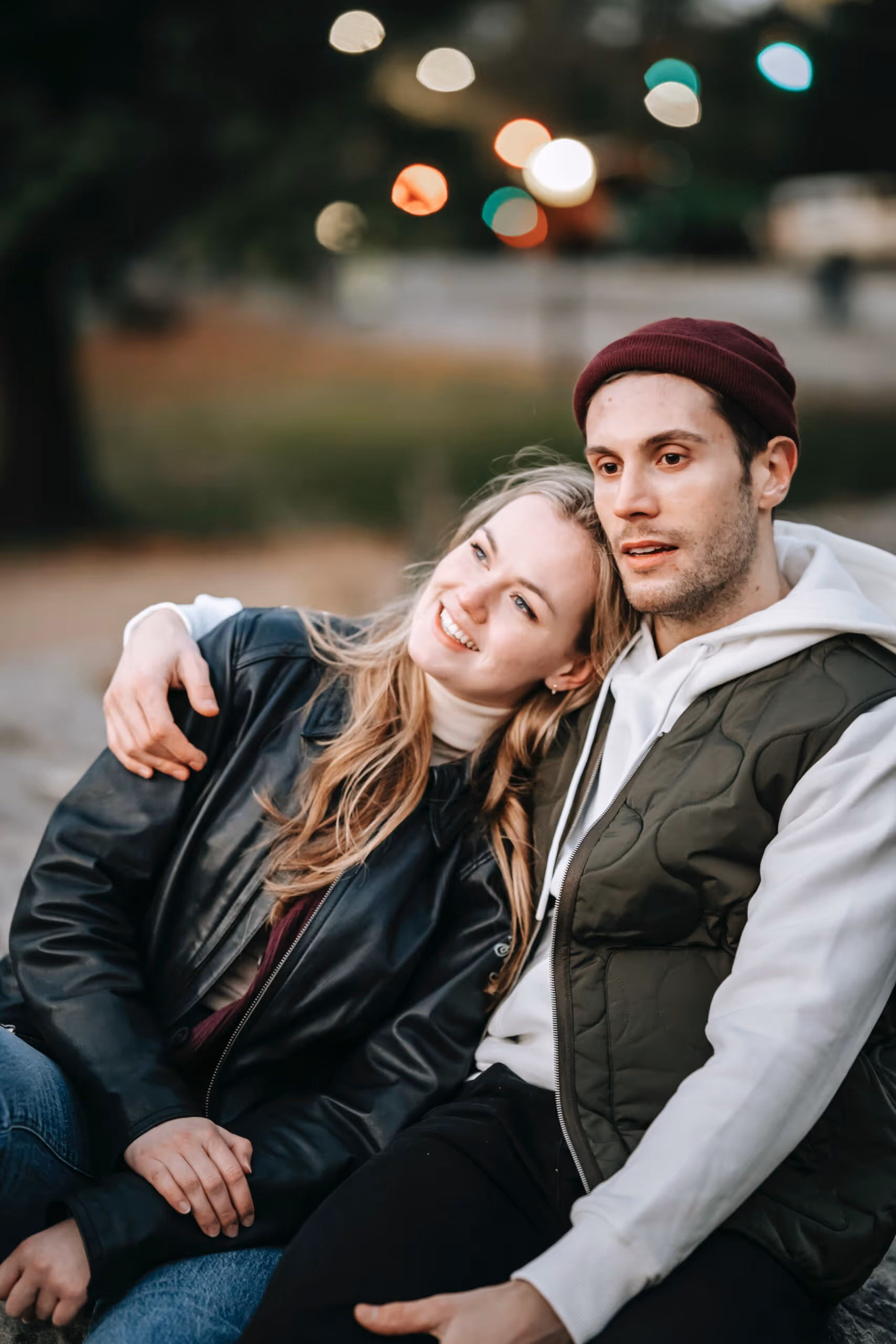 Smiling young couple sitting close together outdoors at dusk, woman leaning her head on the man's shoulder.