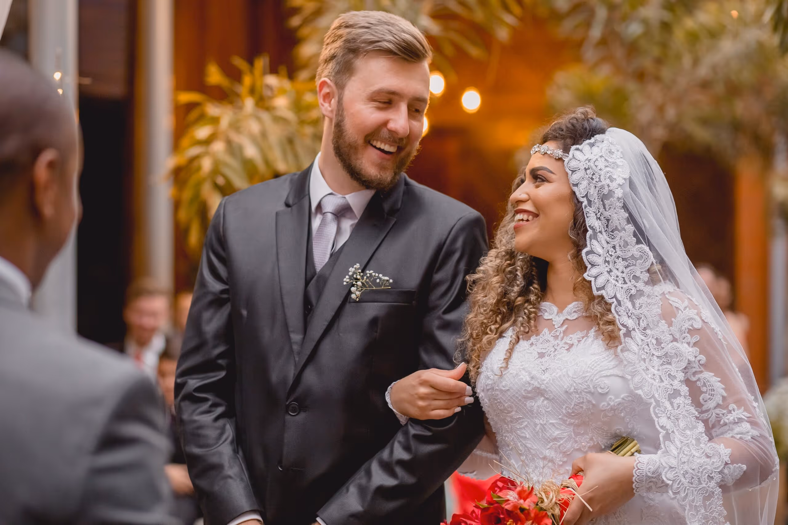 Bride in white lace dress and veil holding red bouquet, smiling and linking arms with groom in black suit during wedding ceremony.