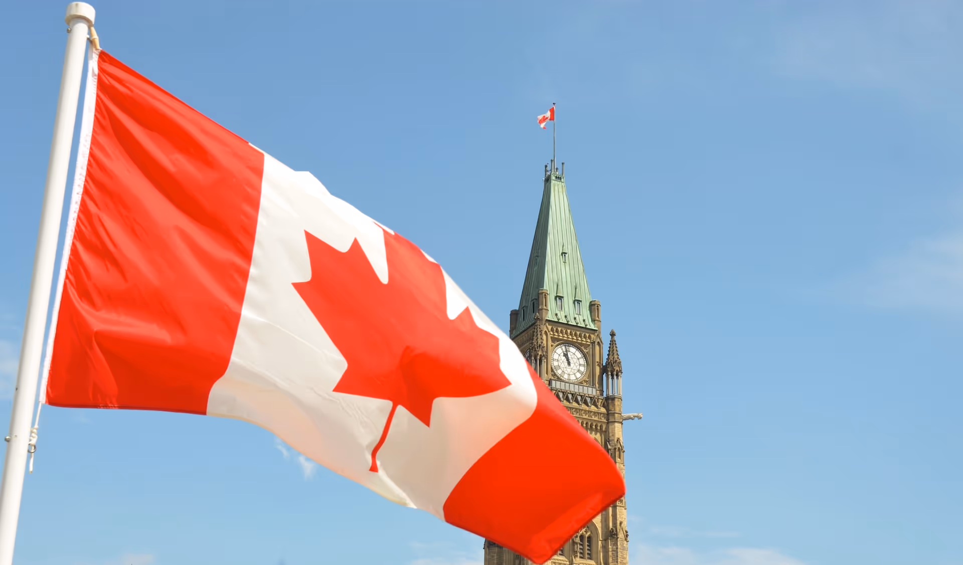 Canadian flag waving in front of the Peace Tower clock on Parliament Hill under a clear blue sky.