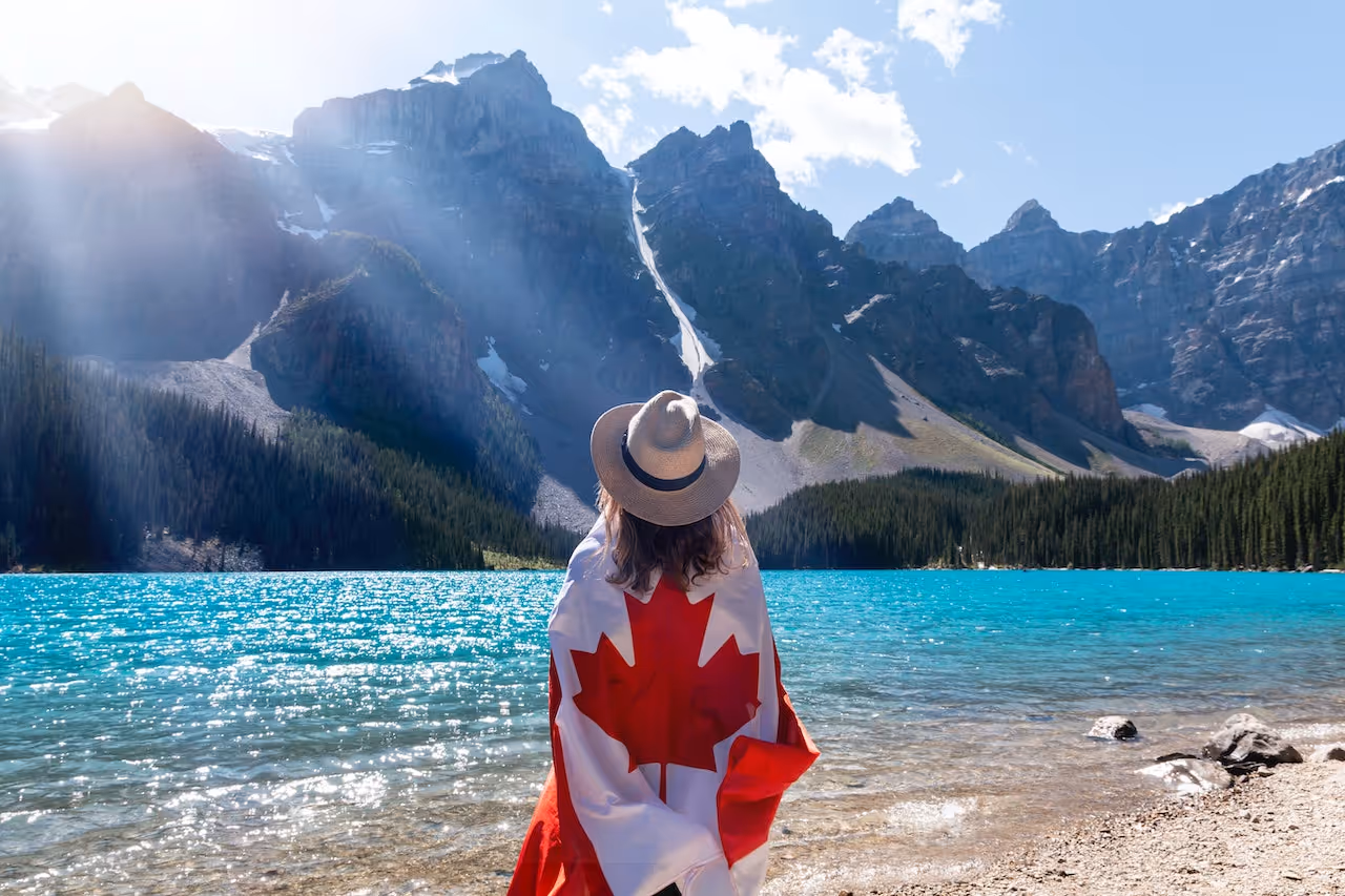 Person wearing a Canadian flag and a hat standing by a turquoise lake with towering mountains in the background.