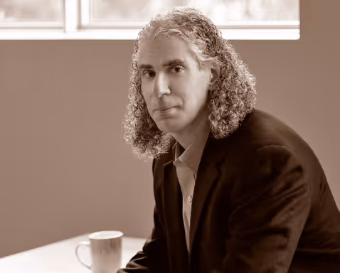 Man with curly hair wearing a dark blazer sitting near a window with a coffee mug on the table.