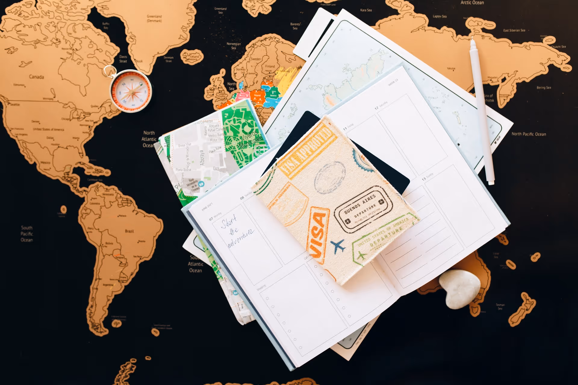 Travel planner setup including open notebooks, a map, a compass, a white pen, and a fabric passport holder on a world map background.
