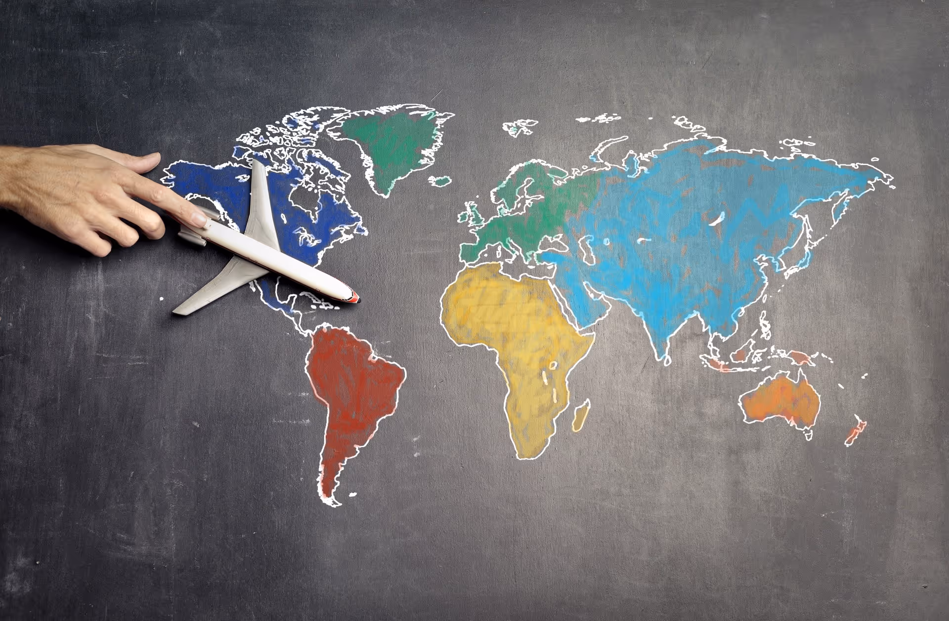 Hand holding a toy airplane over a colorful chalk-drawn world map on a blackboard.