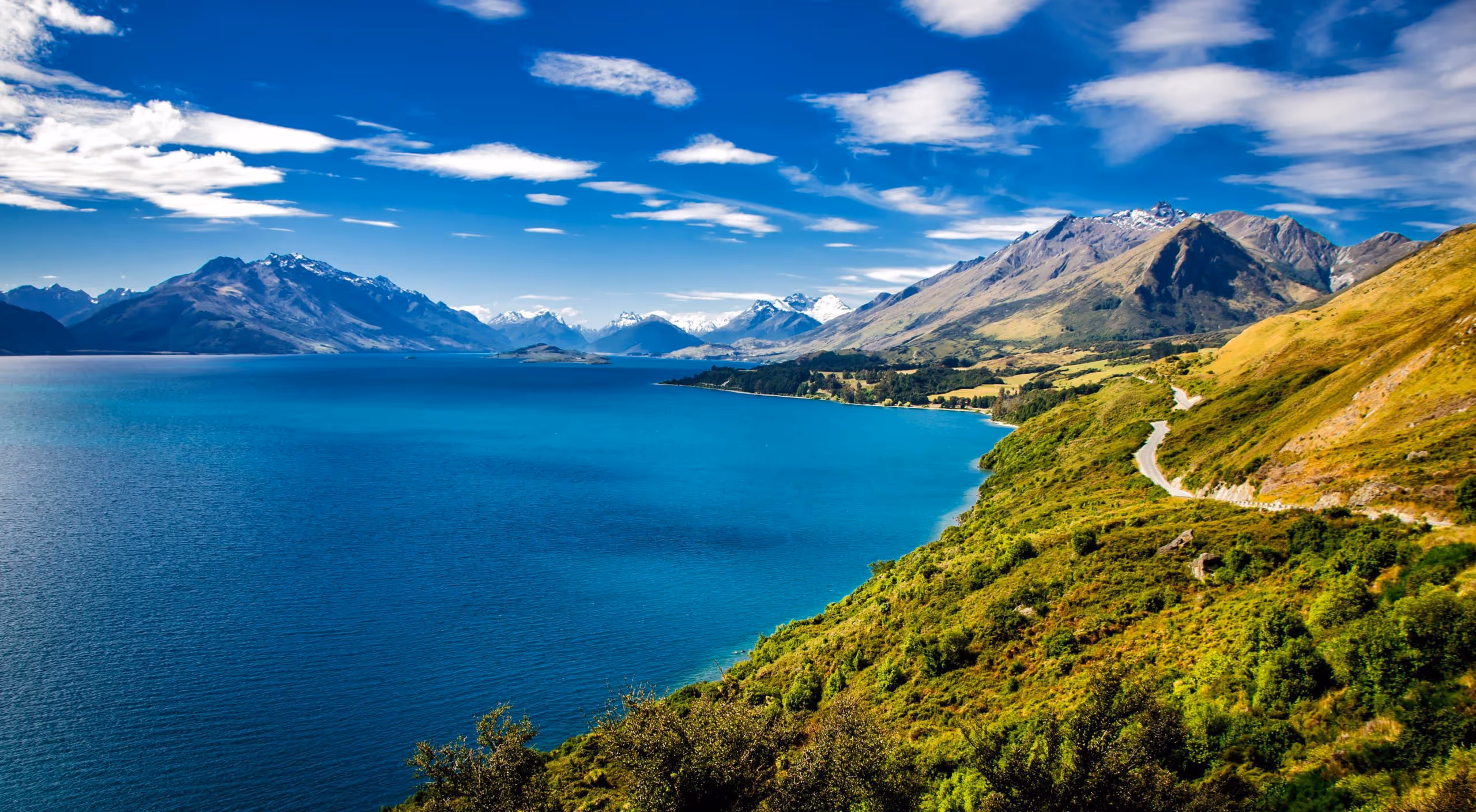 Clear blue lake bordered by green hills and winding road with snow-capped mountains under a blue sky with scattered clouds.