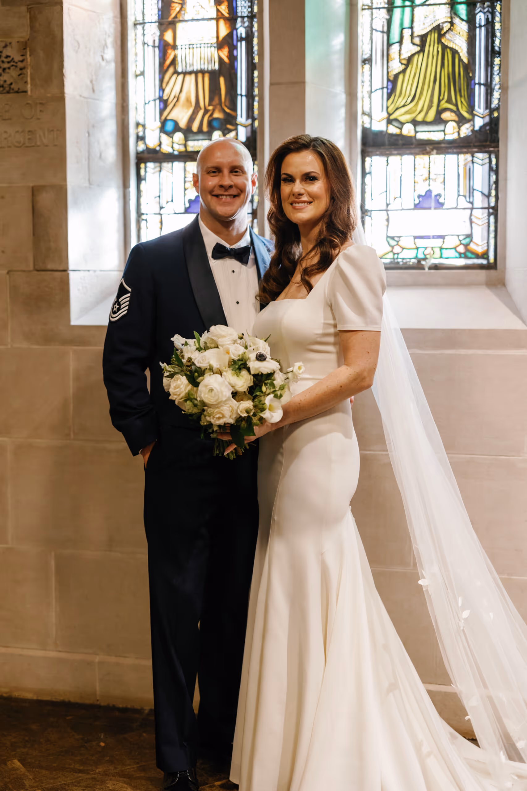 Bride in white gown holding bouquet stands beside groom in formal military uniform inside a church with stained glass windows.