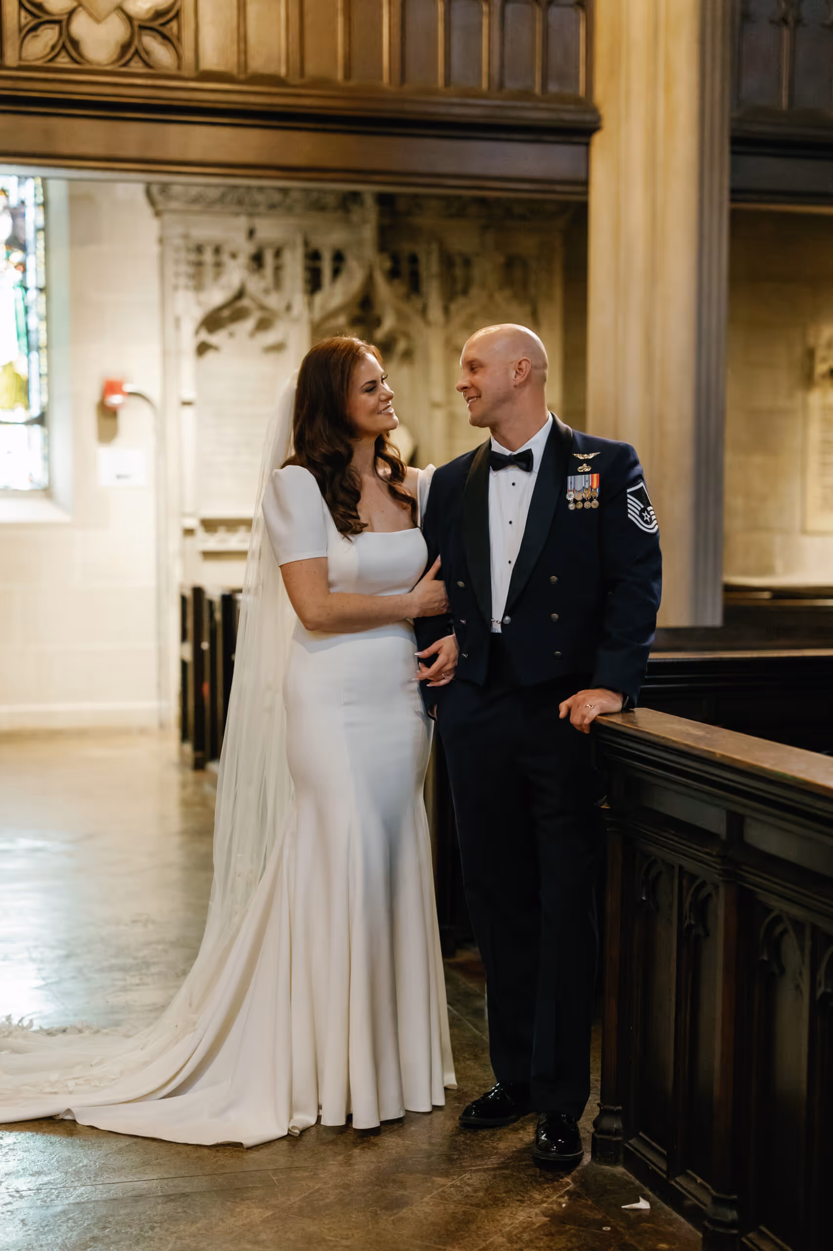 Bride in white wedding gown and veil smiling at groom in military dress uniform inside a church.