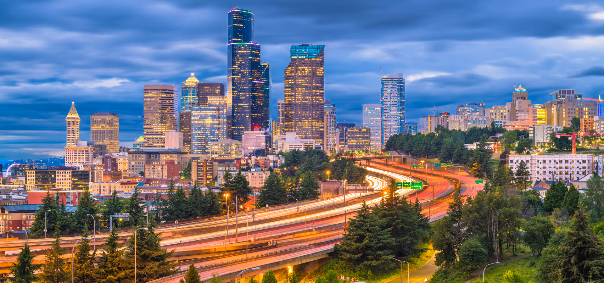 Twilight view of a city skyline with illuminated high-rise buildings and busy highway with light trails surrounded by trees.