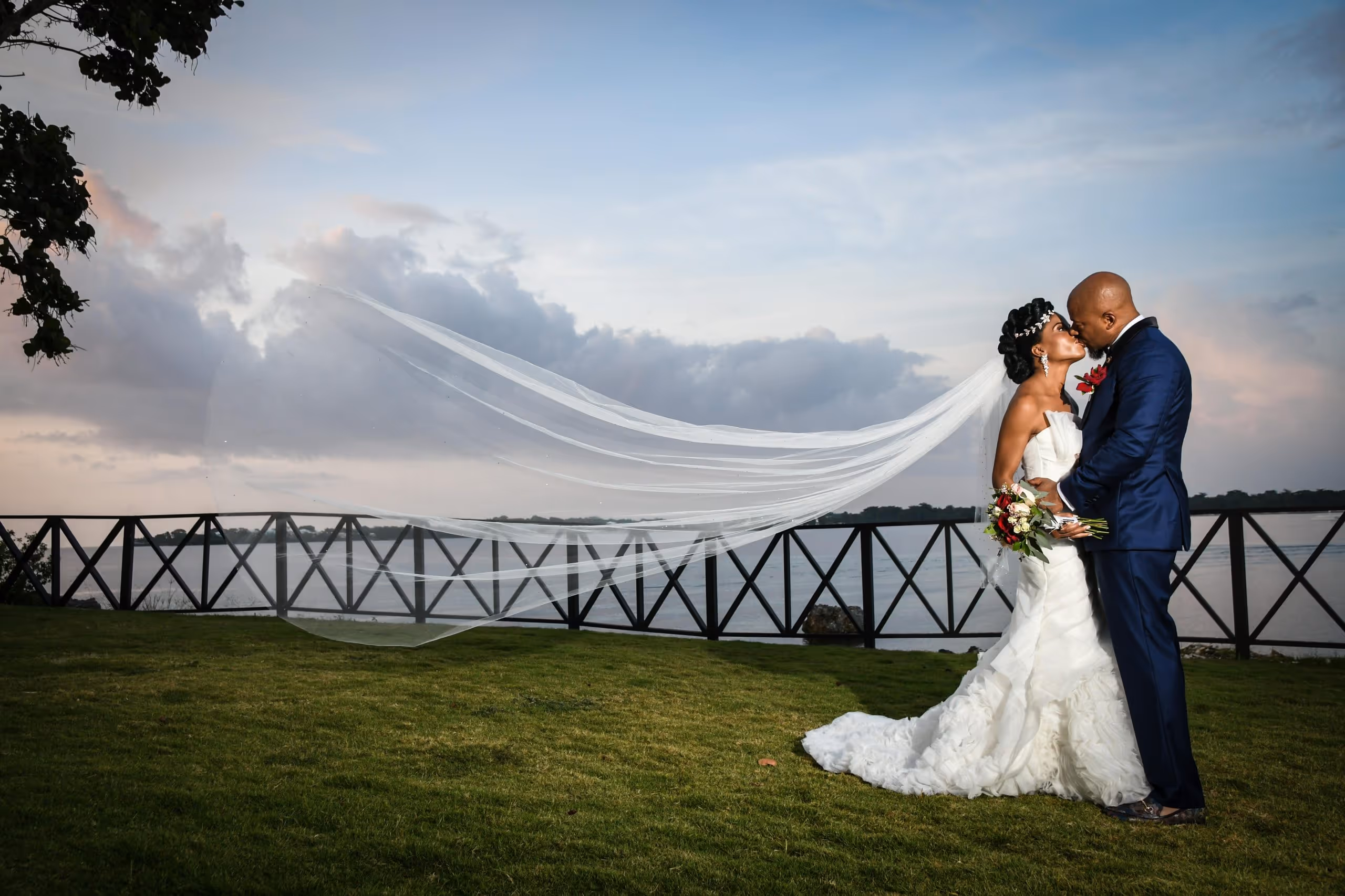 Bride and groom kissing outdoors by a waterfront with the bride's long veil flowing in the breeze.