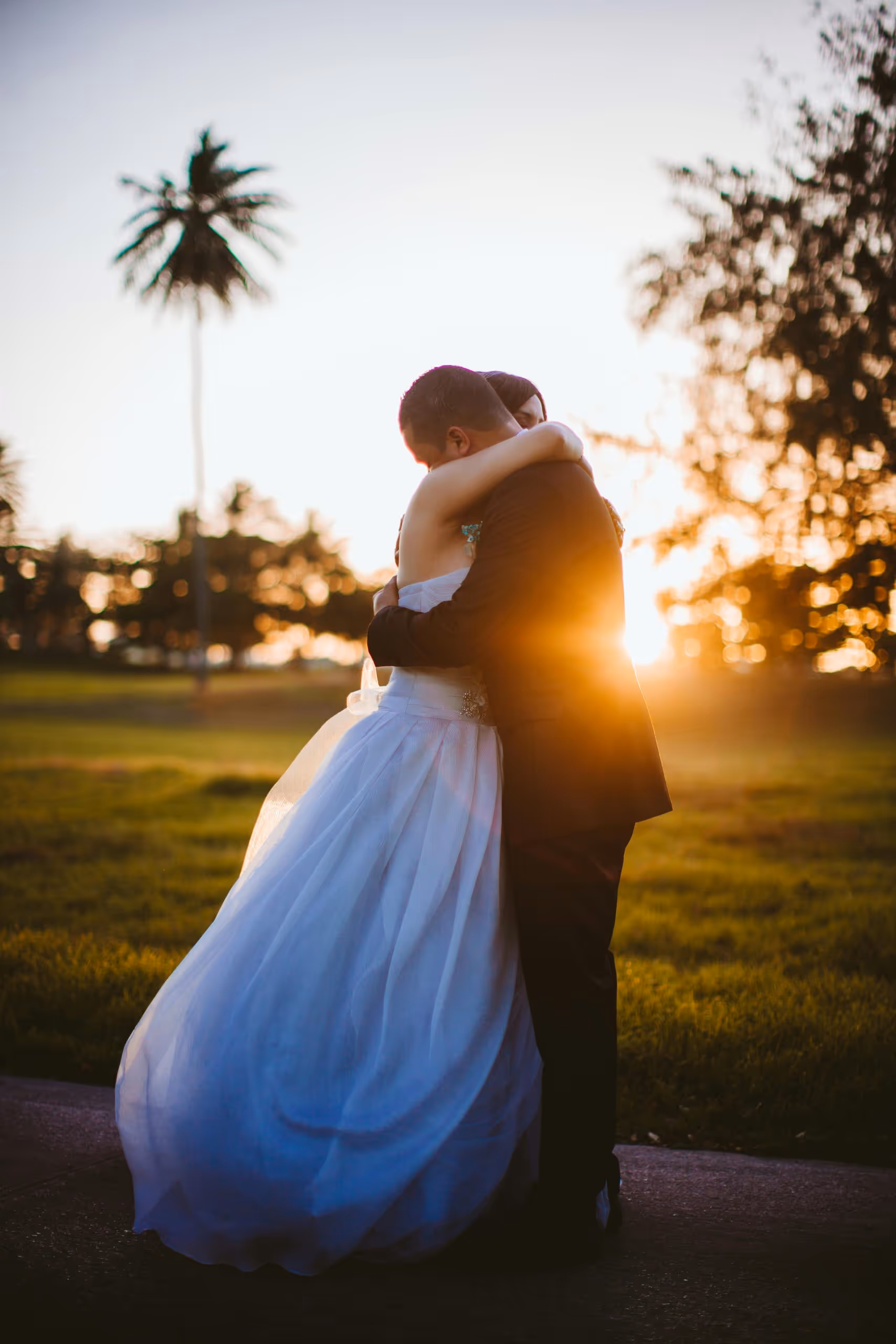 Bride and groom embracing outdoors at sunset with a palm tree and greenery in the background.