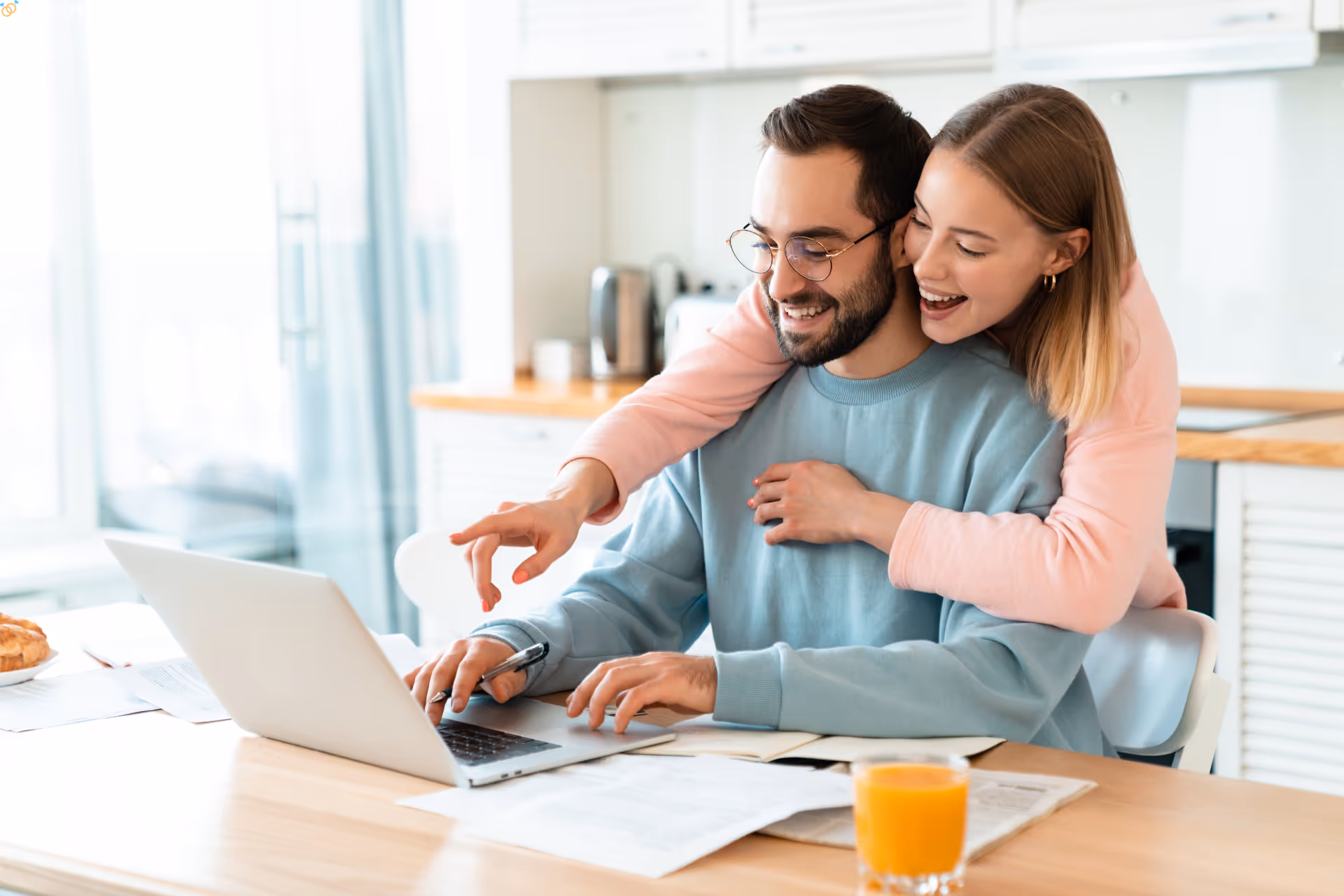 Smiling woman hugging a man from behind as they look at a laptop together in a bright kitchen.