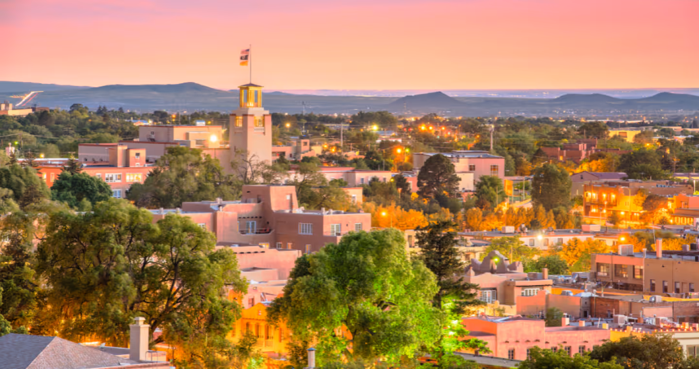 Sunset view over a city with adobe-style buildings, a central tower with a flag, and surrounding trees.