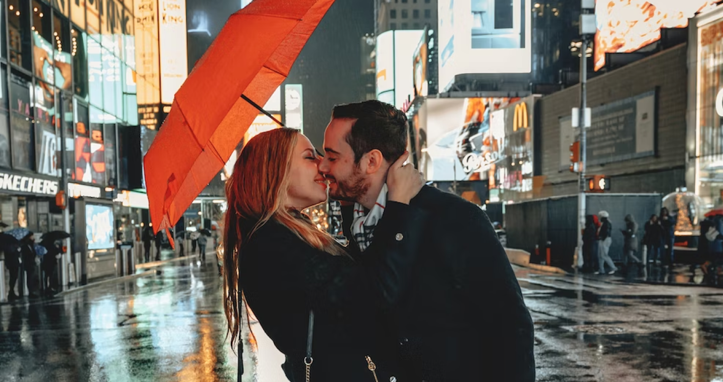 Couple embracing under a red umbrella on a rainy city street at night with wet pavement reflecting bright lights.
