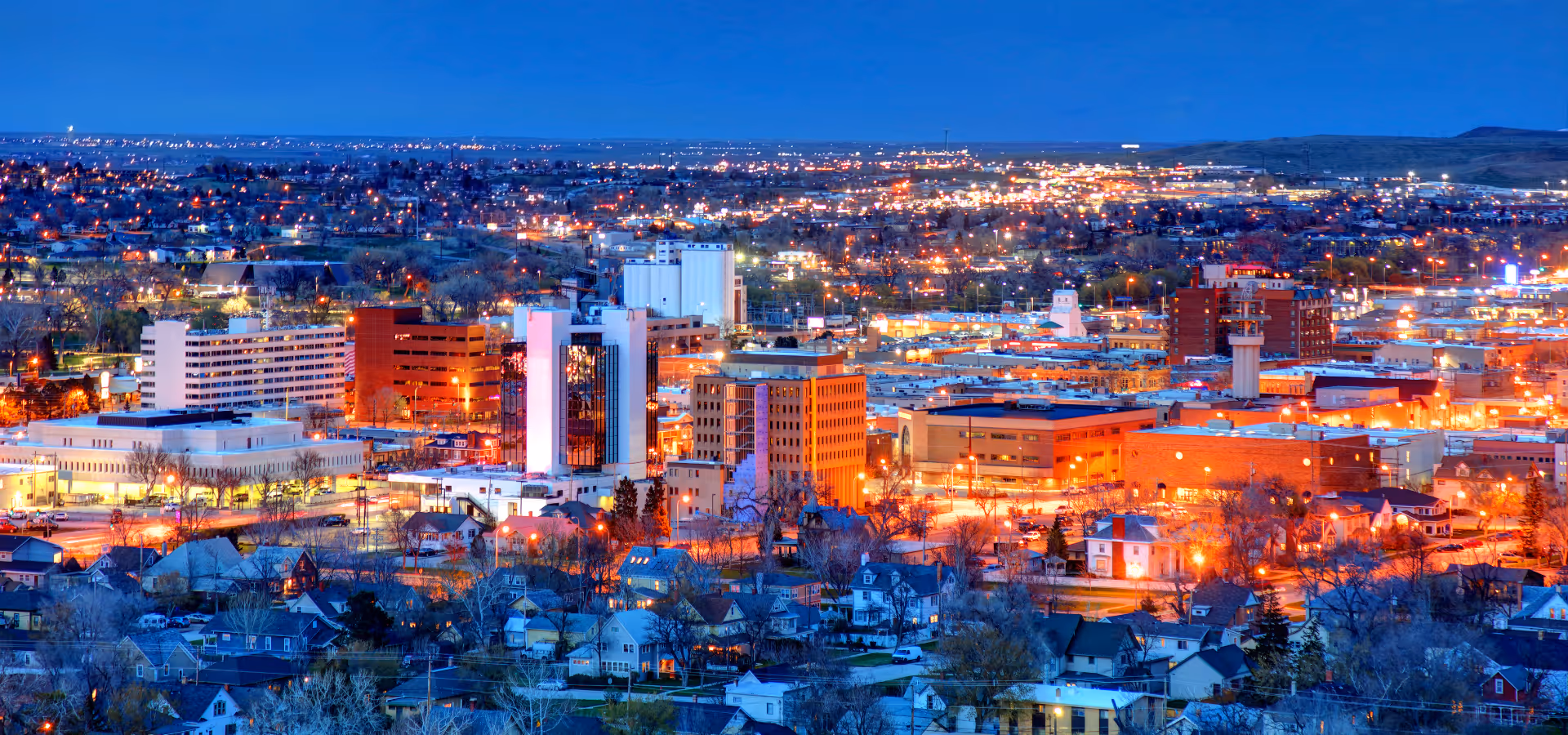 Nighttime cityscape with illuminated buildings and houses under a clear blue sky.