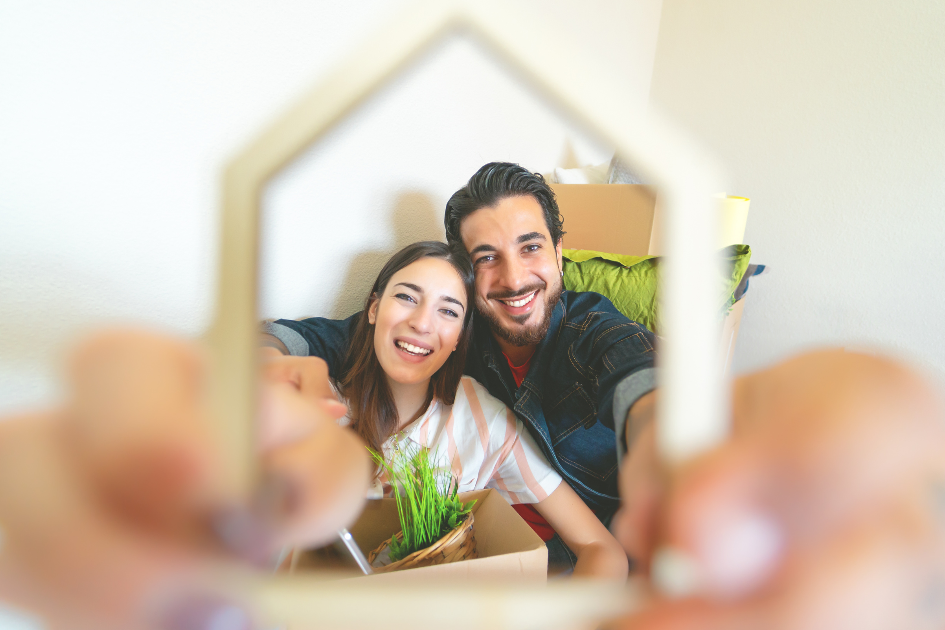 Smiling couple sitting behind a small wooden house frame with moving boxes and a potted plant.
