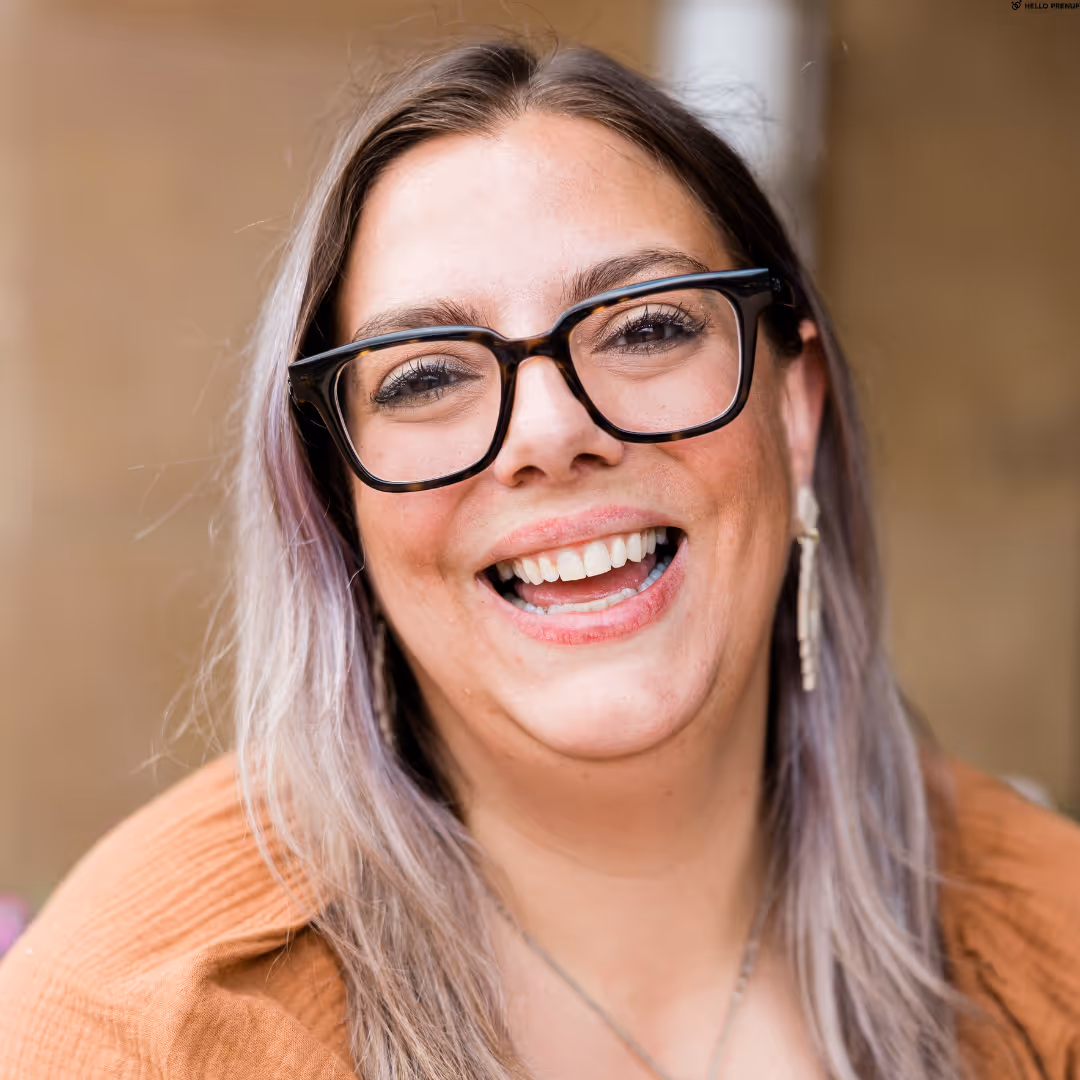 Smiling woman with long light purple hair wearing large black glasses and a rust-colored top.