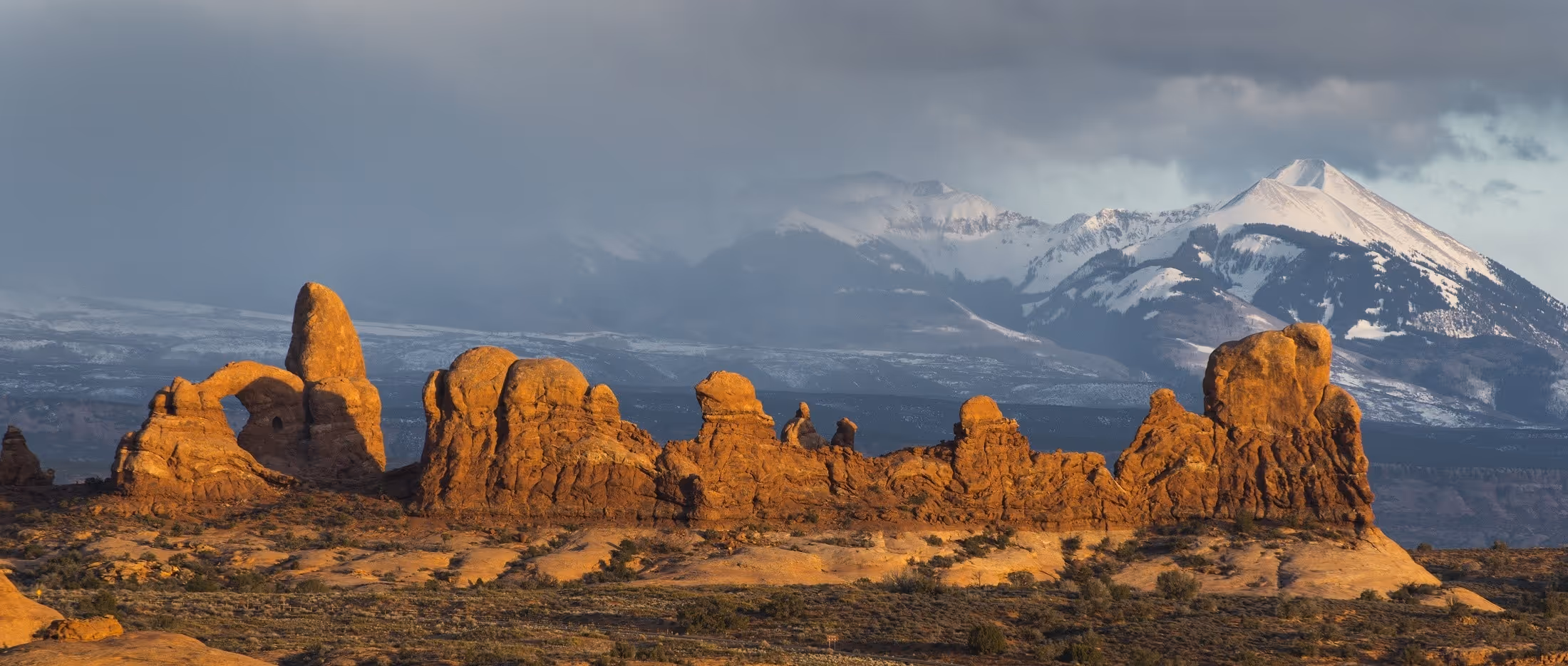 Sunlit orange rock formations in desert with snow-capped mountains under a cloudy sky in the background.