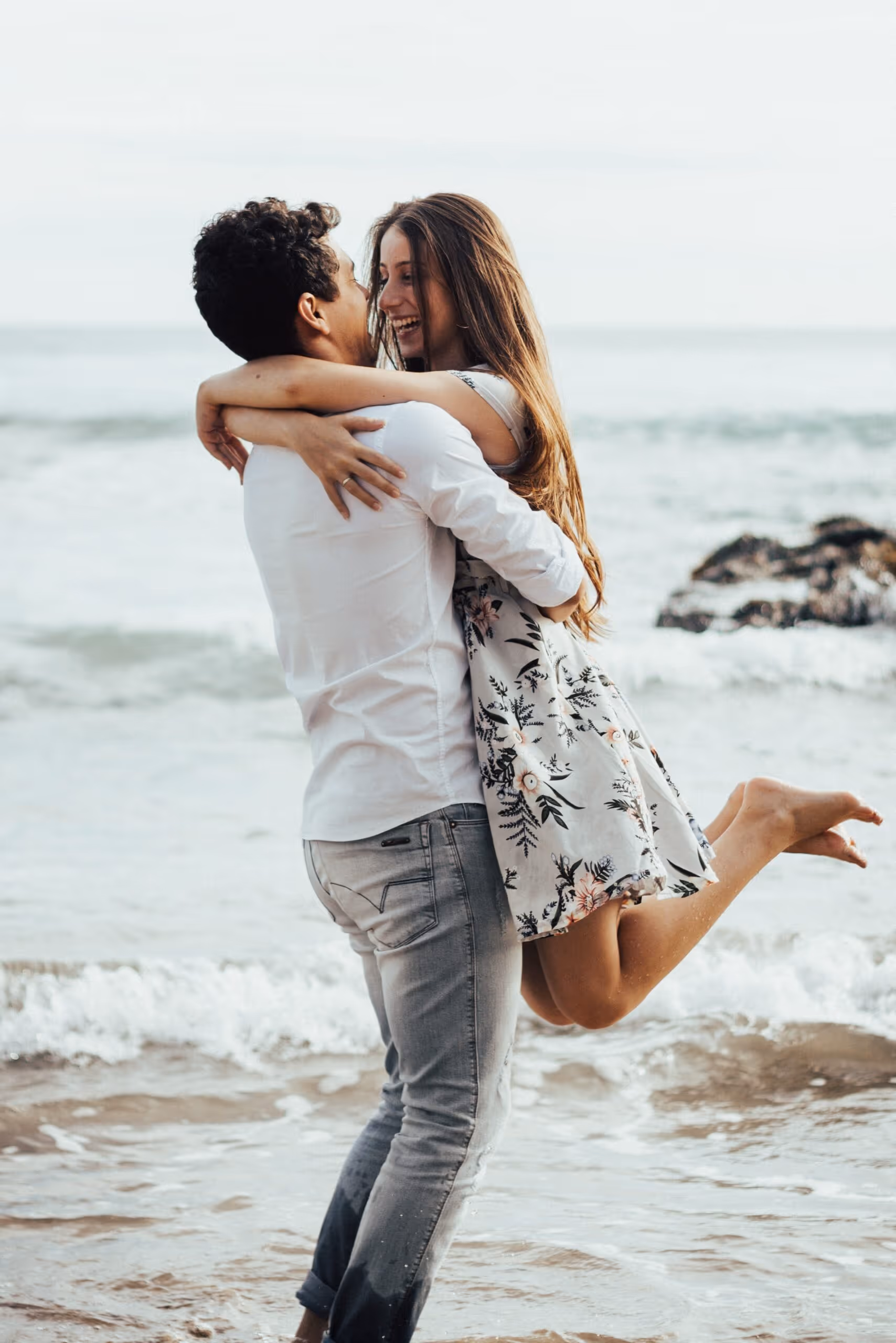Couple embracing and smiling on a beach with ocean waves in the background.