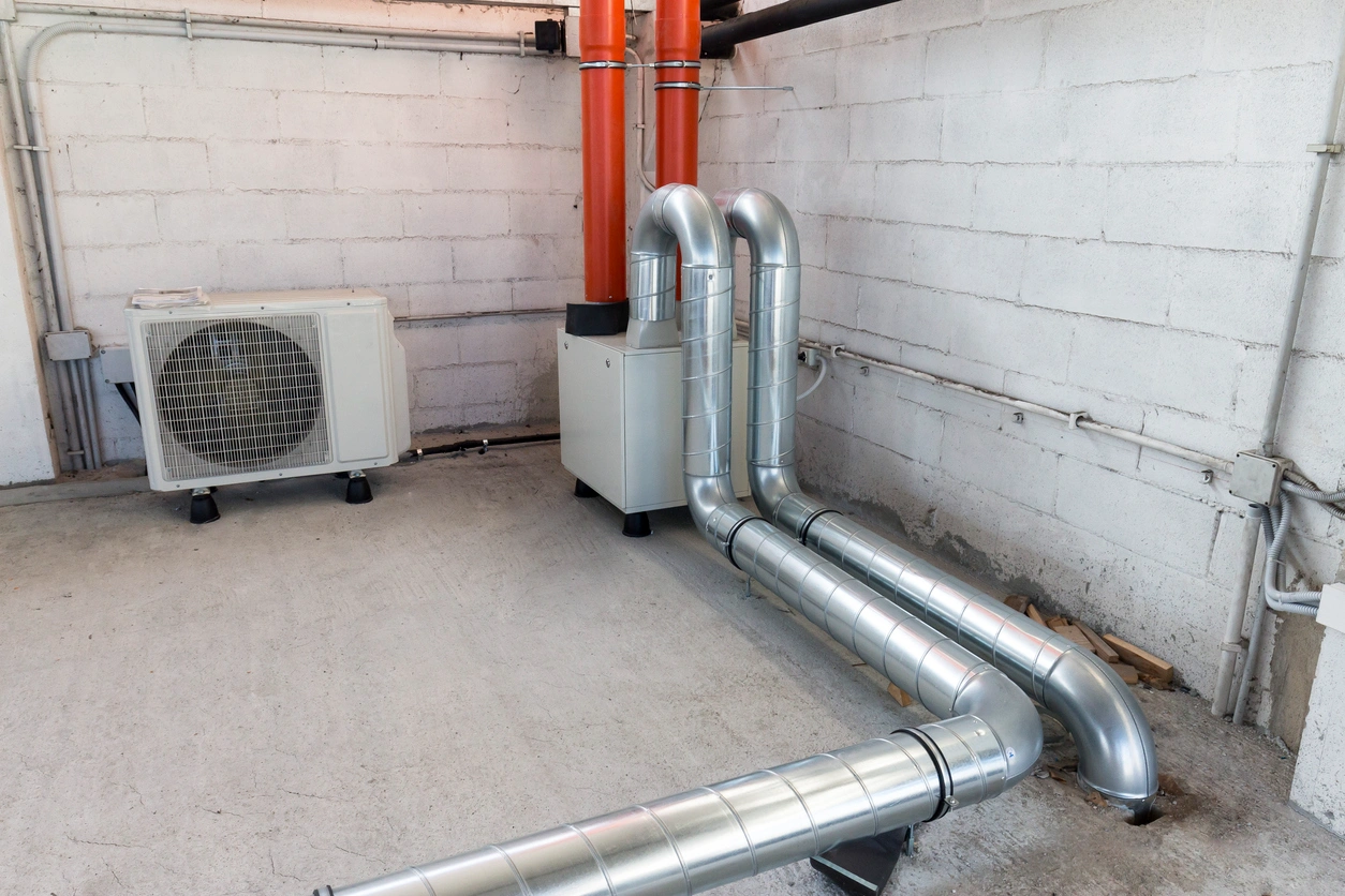 A technical HVAC setup in a basement with silver duct pipes and orange vertical ducts on a white wall.