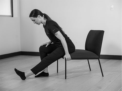 Woman tit closer to the edge of the chair. With one foot flat on the floor, extend the other out straight with the heel on the ground.