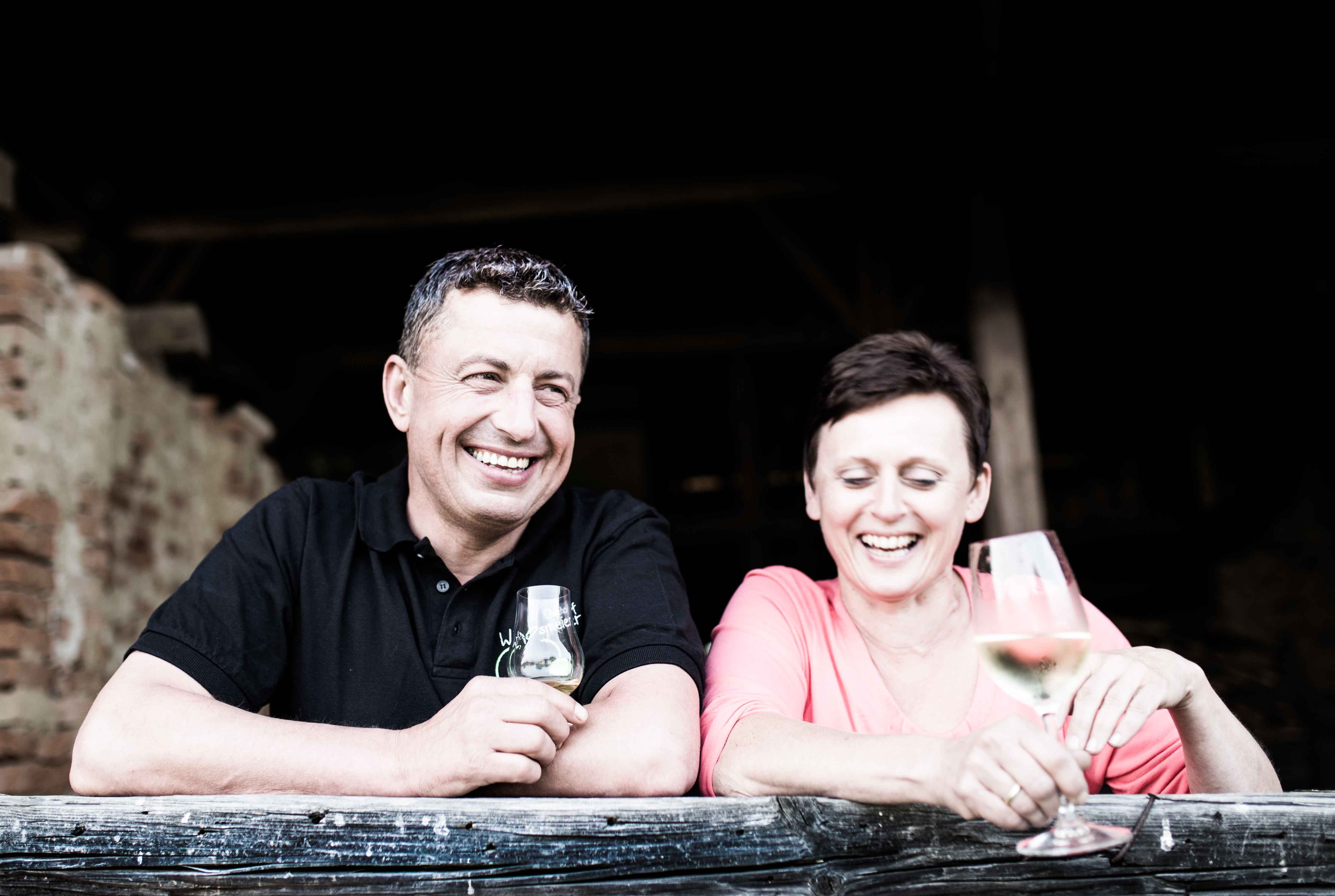 Ein lächelnder Mann im schwarzen Poloshirt und eine lachende Frau in rosa Oberteil lehnen an einem alten Holzzaun und halten jeweils ein Glas Wein.