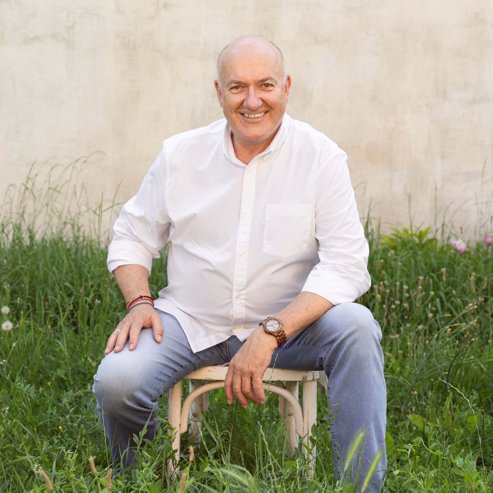 Fröhlicher älterer Mann mit weißem Hemd und Uhr sitzt auf einem Stuhl im grünen Gras vor einer beige Wand.