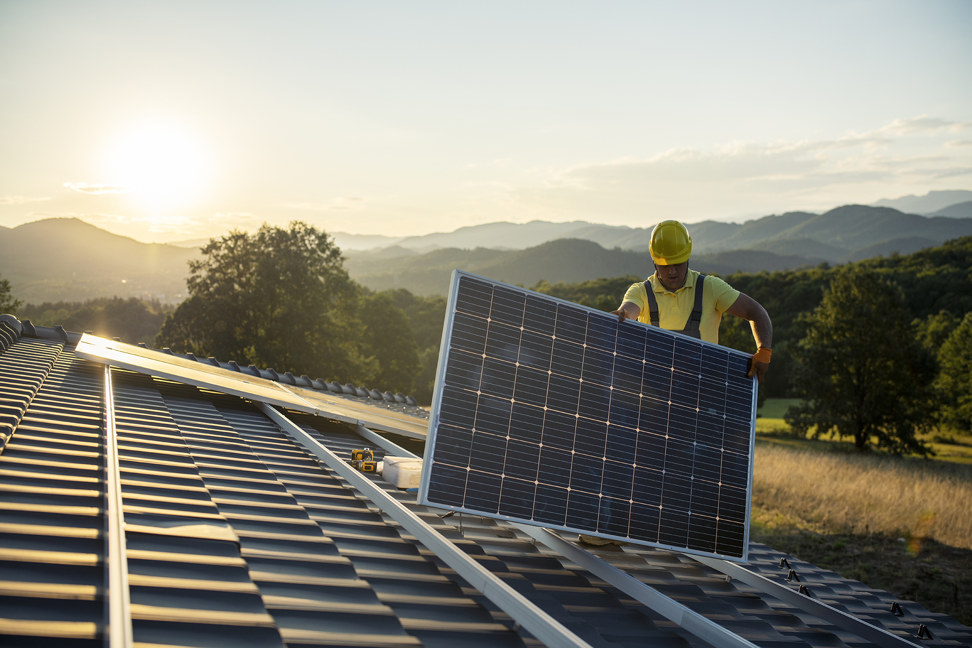 Worker in safety gear installing a solar panel on a tiled roof at sunset with mountains and trees in the background.
