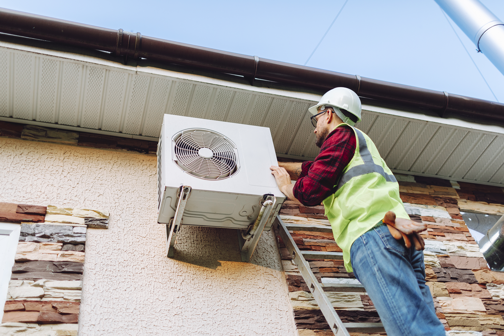 Technician in a safety helmet and vest installing or inspecting an outdoor air conditioning unit on a wall using a ladder.
