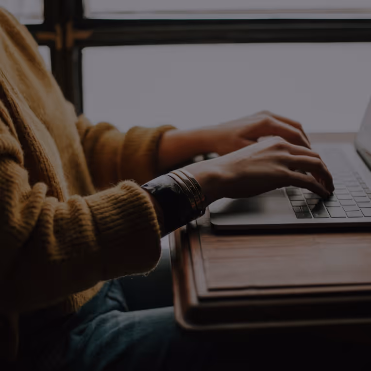 Person wearing a brown sweater typing on a laptop at a wooden desk.
